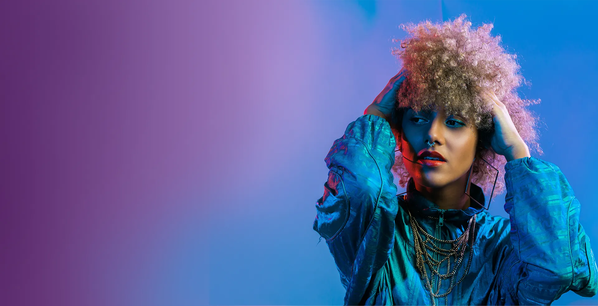 Startseite Teaser Hintergrundbild Braungebrannte Frau mit vielen, lockigen, hochtoupierten, blonden Haaren, die sich in die Haare fast, lassiv zur Seite schaust auf einem Bild mit vielen lila- und blau-Tönen